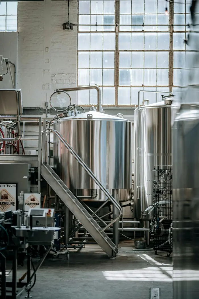 Stainless steel brewing kettles and a steel staircase in a brewhouse lit by tall factory windows.