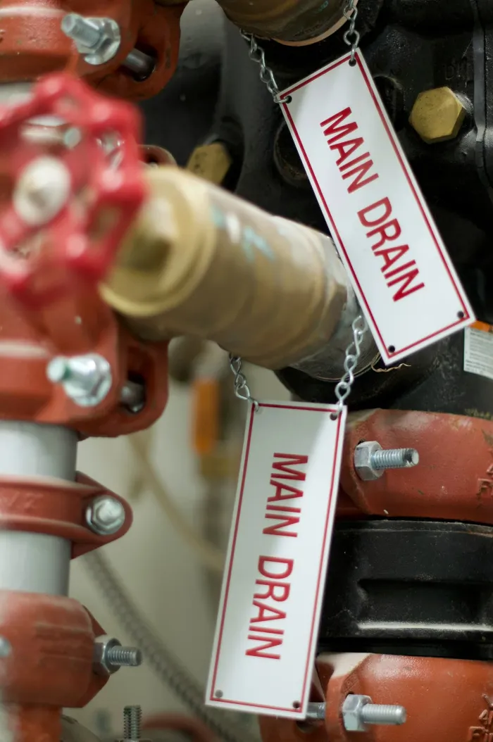 Two white tags reading Main drain hang on chains from a valve and its pipework.